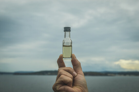 Hand Holding A Small Bottle With Liquid Inside, On A Background Of Sea And Sky