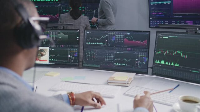 African American trader with pen in hands works at computer with displayed real-time stocks. Colleagues analyze exchange market charts on big screens at background. Cryptocurrency trading. Close up.