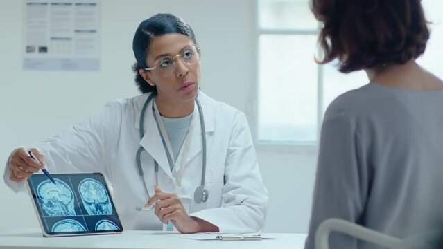 Young African-American female doctor pointing at brain MRI scan on digital tablet, explaining diagnosis to patient, giving health consultation in hospital