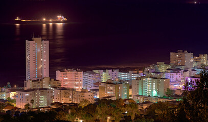 Lights, buildings and city at night with a harbour for urban development, sea water and streets. Dark, travel and aerial view of outdoor residential architecture of downtown with an ocean in evening.
