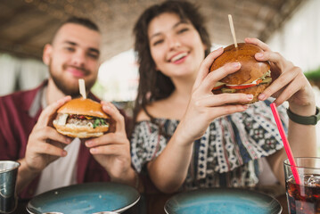 A young couple sits on the summer terrace of a cafe and eats burgers with cola. Man and woman have a great time and relax enjoy food in cafe