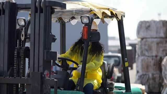 Black-haired worker driving excavator for sorting trash at plant. Long-haired woman wearing hardhat controls process at waste sorting factory