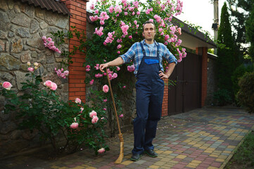 Fototapeta na wymiar Full length portrait young adult handsome Hispanic male gardener, sweeper with a broom smiling, looking confidently at camera. People. Professional occupation. Mansion cleaning and maintenance concept