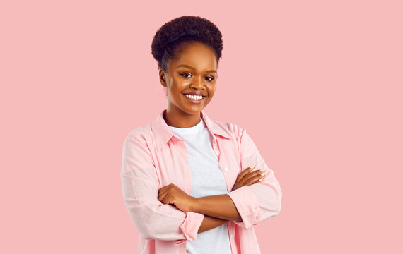 Studio portrait of happy woman. Confident attractive young African American lady in casual pink shirt and white tee standing with her arms folded on pink background, looking at camera and smiling