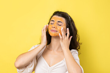 A young brunette girl with a cleansing scrub mask on her face, looks at the camera and smiles, against a yellow background.