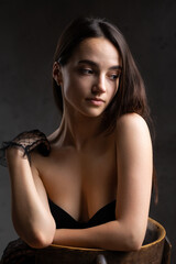 Classic dark studio portrait of a young brunette woman in black clothes who is sitting on a chair.