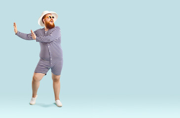 Funny eccentric fat man in good mood dancing and fooling around enjoying summer vacation. Bearded overweight man in striped swimwear, sunglasses and beach hat on pastel light blue background. Banner.