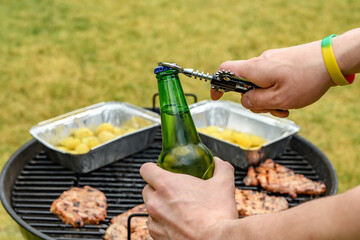 Zielona butelka z piwem otwierana otwieraczem w ogrodzie na tle grilla z kawałkami mięsa wieprzowego - obrazy, fototapety, plakaty