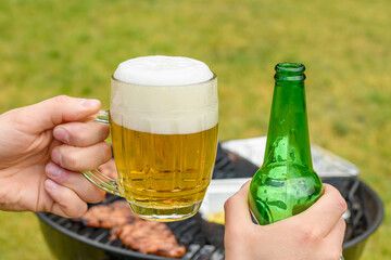 Trzymany w dłoni kufel z jasnym piwem i zielona butelka, impreza z alkoholem w plenerze