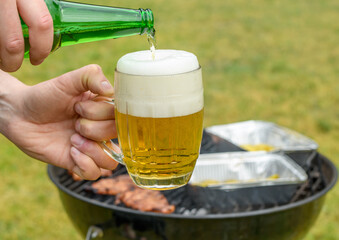 Piwo jasne nalewane z butelki do kufla na tle rozpalonego grilla pełnego jedzenia