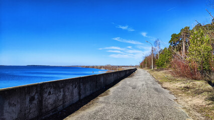 Fototapeta na wymiar Asphalt path on the embankment near the water of the lake, sea, river, reservoir and blue distance with the sky with white clouds and horizon. Natural landscape with water on a sunny day
