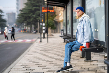 Young blond guy student waiting transport very long time in morning.Man on bus stop with coffee...