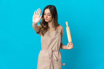 Young Romanian woman holding a rolling pin isolated on blue background making stop gesture and...
