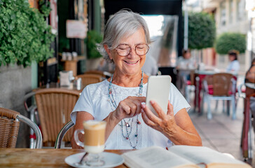 Attractive relaxed senior woman sitting at outdoor cafe with coffee and book while using phone to read a message, smiling elderly enjoying retirement and free time