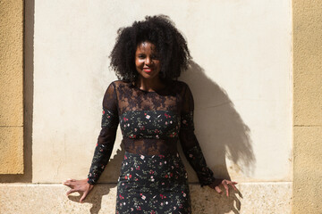 Portrait of black woman, young and beautiful with afro hair and black dress with flowers is very happy and smiling. In the background a light brown wall. Concept different expressions.