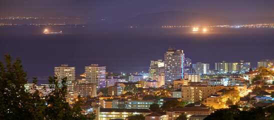Obraz premium Harbour, buildings and city at night with lights, urban development and streets. Dark, travel and outdoor residential architecture of downtown with ocean or sea water in the evening with a skyline.