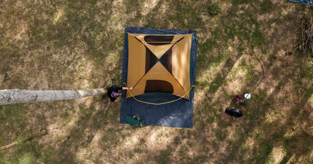 Top view of Asian male tourist pitching a tent under a tall pine tree.