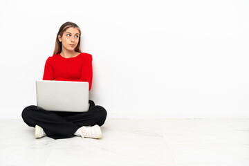 Young caucasian woman with a laptop sitting on the floor making doubts gesture while lifting the shoulders