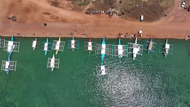 Aerial Drone Bird's Eye Top Down View of multiple Filipino Outrigger Bangka Boats moored to a quaint dock, revealing tropical village town and settlers inland. Vacation shot from the Philippines