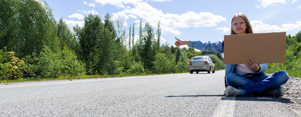 A young girl is sitting on the road with a sign in her hands, hitchhiking around the country. The...