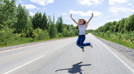 Fototapeta premium A smiling young girl, a teenager, jumps in the middle of the road in the summer outside the city. The concept of tourism and travel advertising. High quality photo