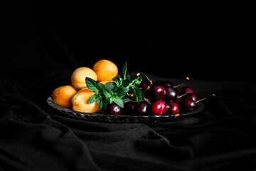 ripe yellow apricots and dark cherries lie on a glass plate with mint sprigs on a black background. summer food. the right snack. healthy food for vegetarians