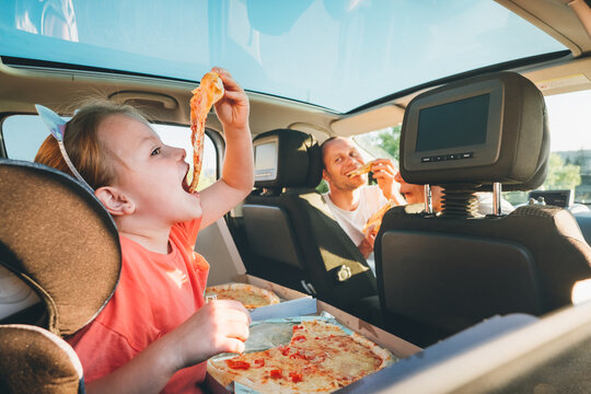 Little girl with open mouth portrait eating Italian pizza sitting in modern car with mother and father. Happy family moments, childhood, fast food eating or auto journey lunch break concept image..