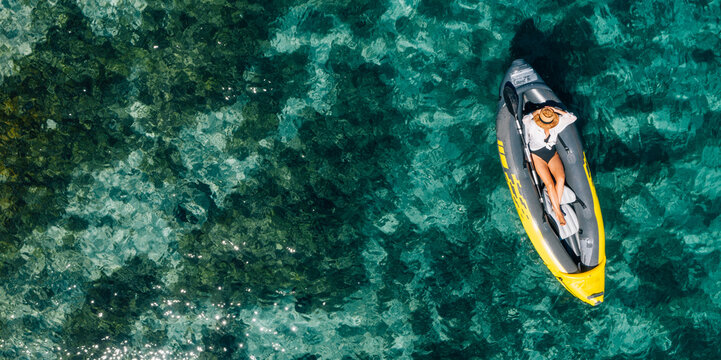 A lonely female in a straw hat smiling, relaxing lying floating in a kayak on the turquoise Adriatic Sea waves. Aerial coastal top view shot. Exotic countries vacations concept.