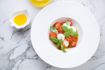 Italian caprese with mozzarella balls, red cherries and green basil served in a white plate, high angle view on a white marble background, horizontal shot