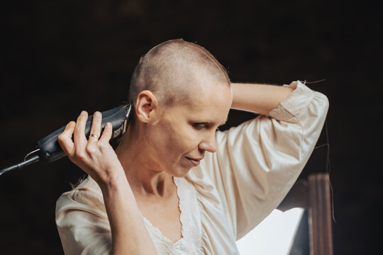Portrait of a bald woman cutting her hair with a clipper on a dark background.