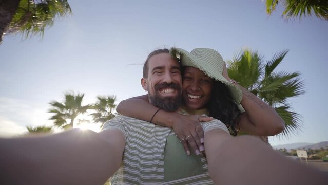 POV of sweet romantic multiracial couple making selfie video kiss with mobile phone on summer beach sunny day. Happy young love people enjoying summer vacation outdoors. Holiday travel and getaways.