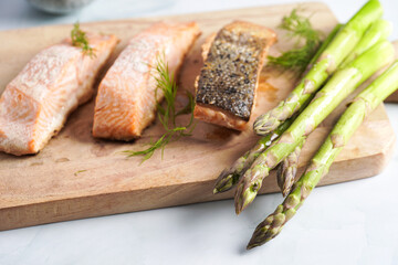 Three grilled fish steaks - red salmon with skin and green asparagus on wooden board, dill, olive oil, avocado on pastel blue background
