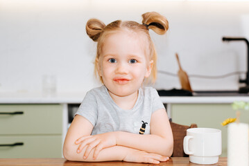 Portrait of single funny child girl sitting in kitchen at table and looking at camera