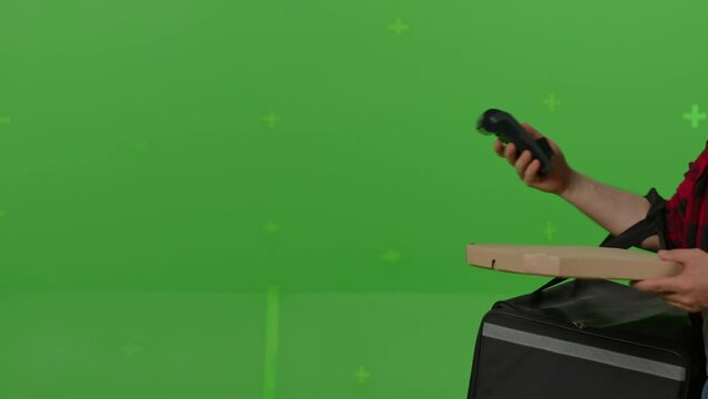 Caucasian male courier passing a box of pizza on green screen chroma key background. Man hold wireless bank payment POS terminal to process acquire credit card payment. Copy space mock up advertising