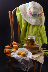 Still life with tea in a teapot, tulips, a hat and lemons on a chair on a black background - 611671137