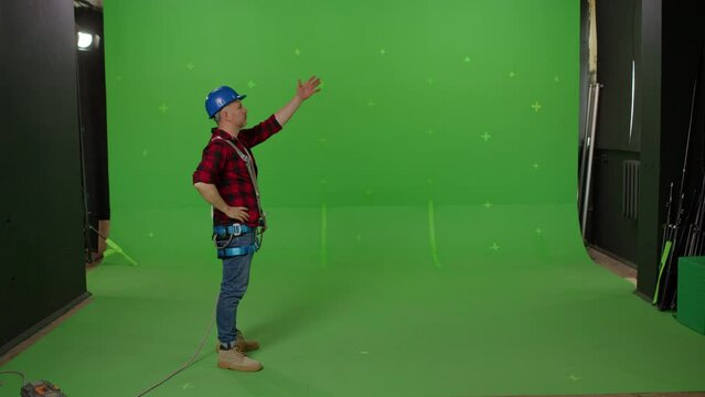 A male builder in professional ammunition and a construction helmet waving his hands and signaling to someone on a green background