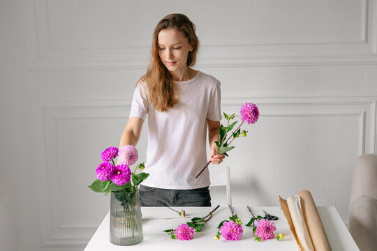 Horizontal image of a young female florist with loose hair picking pink dahlias into a bouquet at the table. Concept of farming, small business