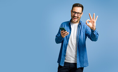 Portrait of satisfied businessman using smart phone and showing OK gesture against blue background - obrazy, fototapety, plakaty