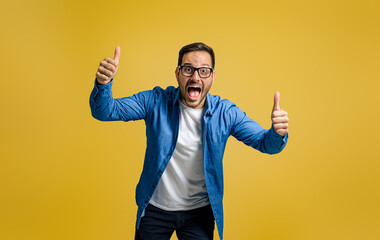 Portrait of exited businessman showing thumbs up and screaming in joy against yellow background