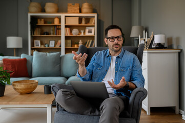 Serious businessman gesturing at camera while working over laptop on armchair in home office