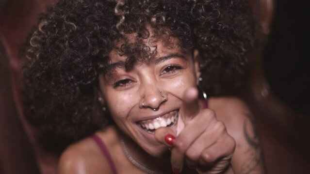 Young afro-haired and tattooed girl sitting in an old-fashioned armchair, smiling and making jokes at the camera. (2/2)