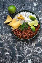 Plate with chili con carne, mixed rice and nachos on a dark-brown granite background, vertical shot with copy space, top view