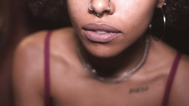 Extreme close-up of a young tattooed afro-haired girl wearing a shiny silver necklace and her luscious lips that seem about to part for a kiss.