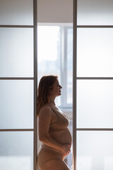 A pregnant woman in nude lingerie stands in profile between folding doors.