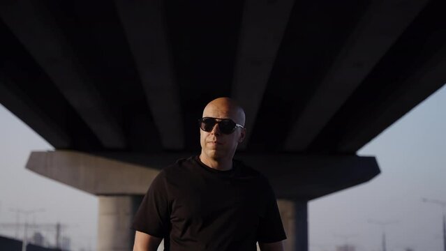 A brutal man walks under the large arch of a road bridge. Stylish biker walks in black glasses and clothes. Modern style of a man. Sunset light on a man without hair.
