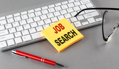 JOB SEARCH text on a sticky with keyboard, pen glasses on grey background