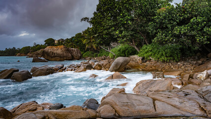 The waves of the turquoise ocean are beating against the coastal granite boulders, foaming. Lush tropical vegetation on the shore. Clouds in the sky. Seychelles. Mahe. Long exposure.