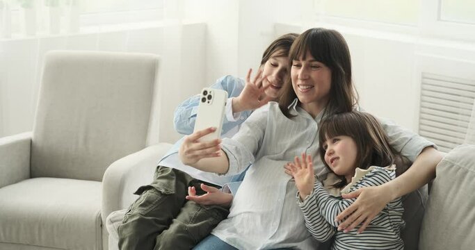 Delightful Caucasian family gathered on the comfortable couch, a pregnant mother, along with her son and daughter, eagerly engage in a video call with beaming smiles on their faces.