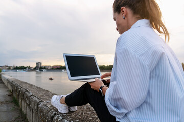 Urban woman sitting on the city waterfront and using her laptop, meeting the end of the work day out of office.