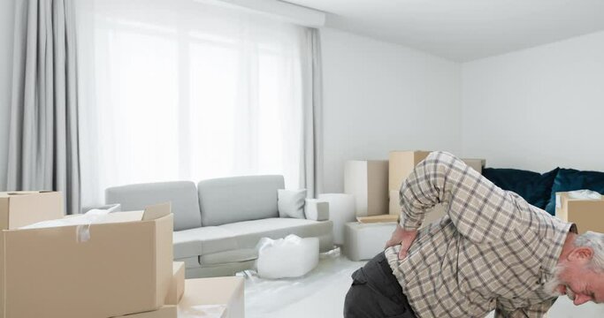 Senior man lifts a heavy moving box and injures his back, experiencing a great deal of pain